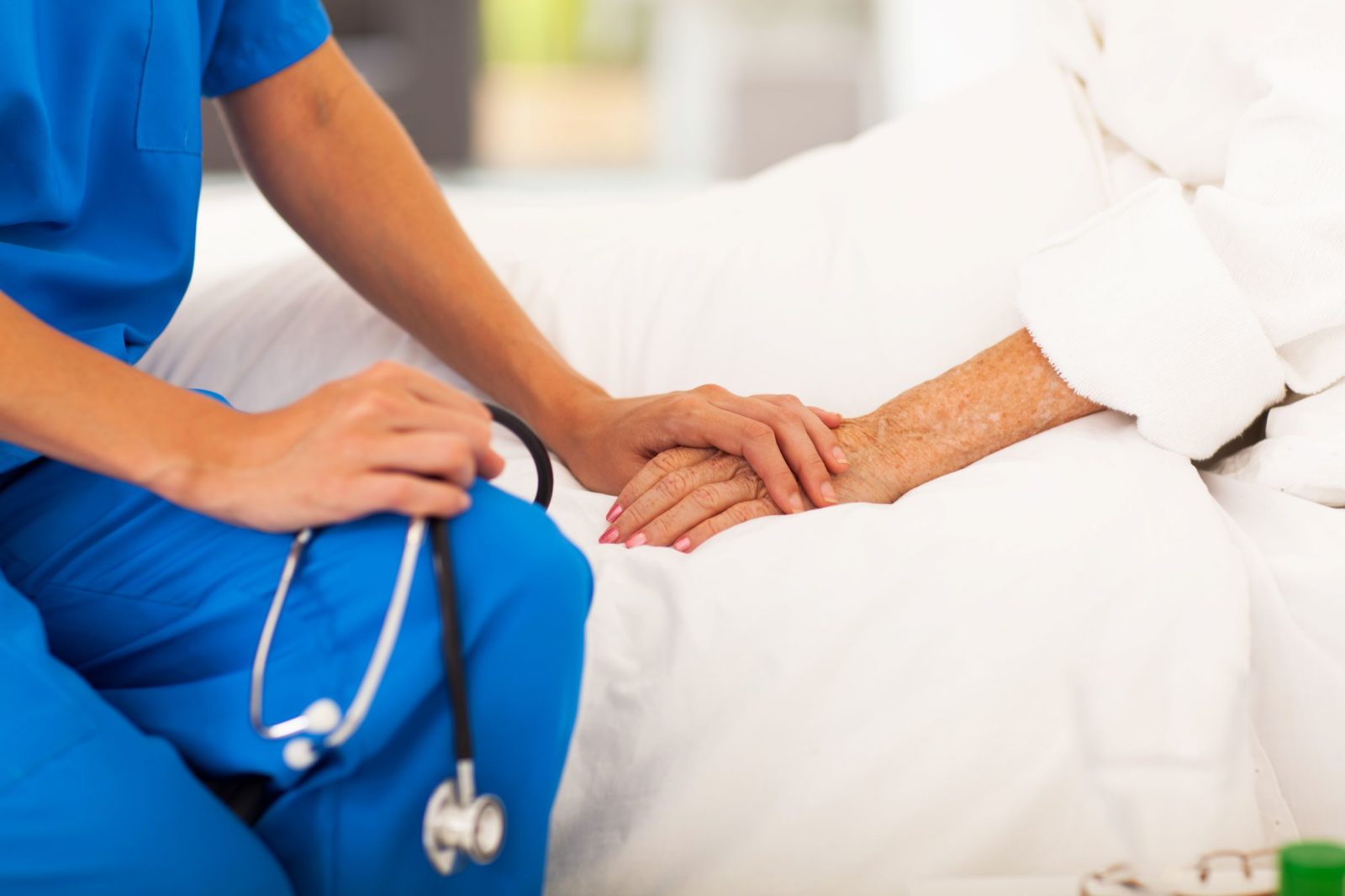 A clinician in blue scrubs holding a patient’s hand at the bedside