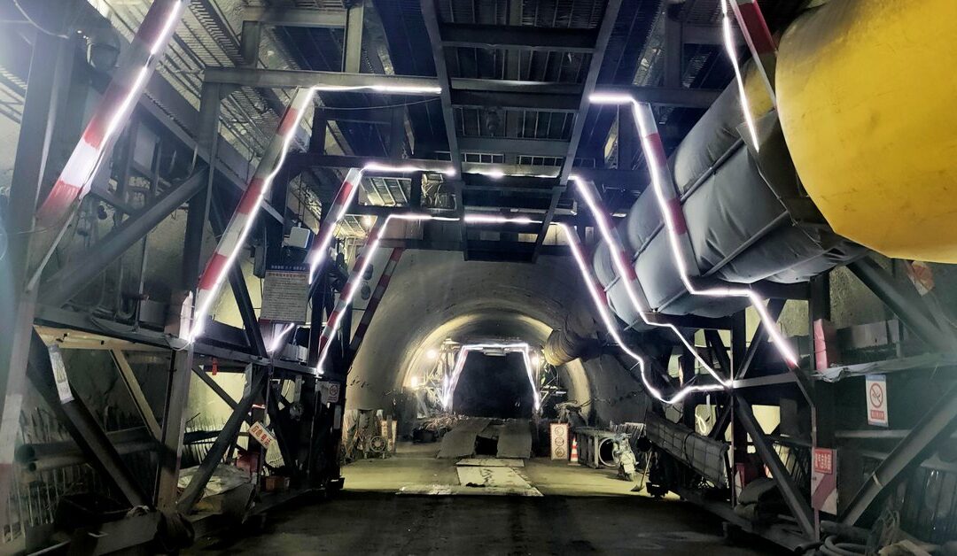 Steel gantry and ventilation ducting inside an unlined highway tunnel bore under construction, lit by LED strips