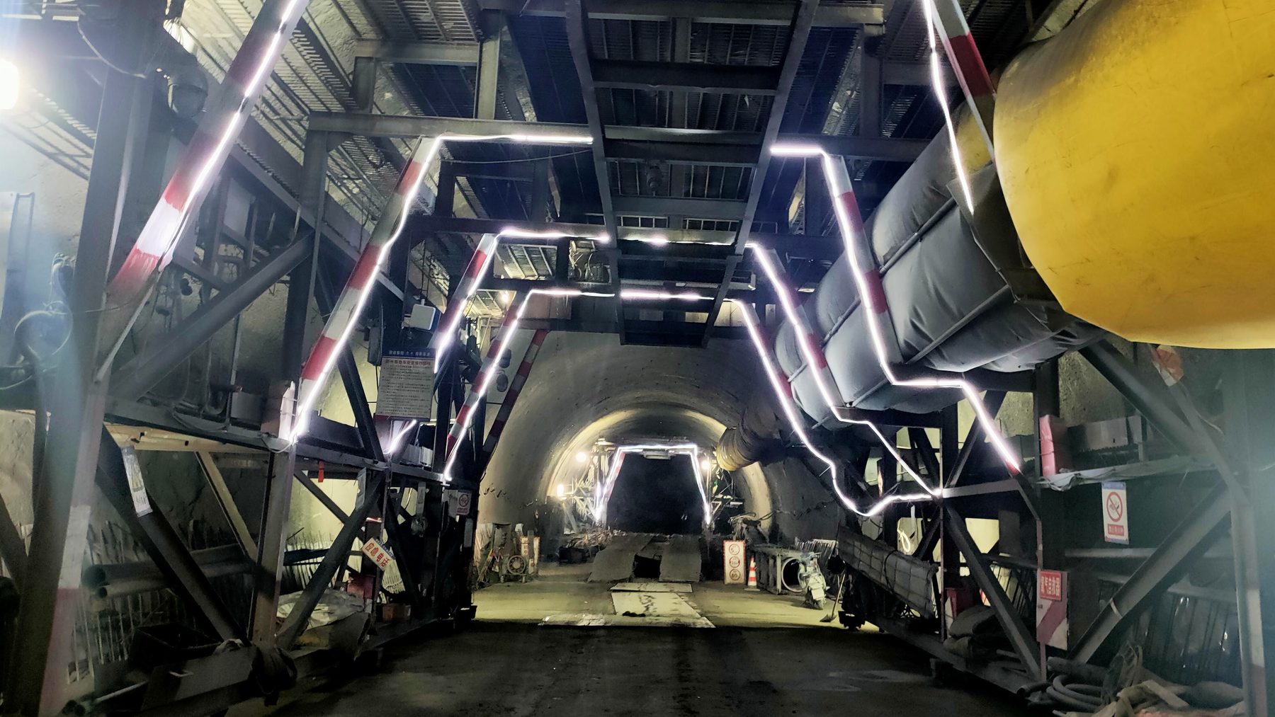 Steel gantry and ventilation ducting inside an unlined highway tunnel bore under construction, lit by LED strips