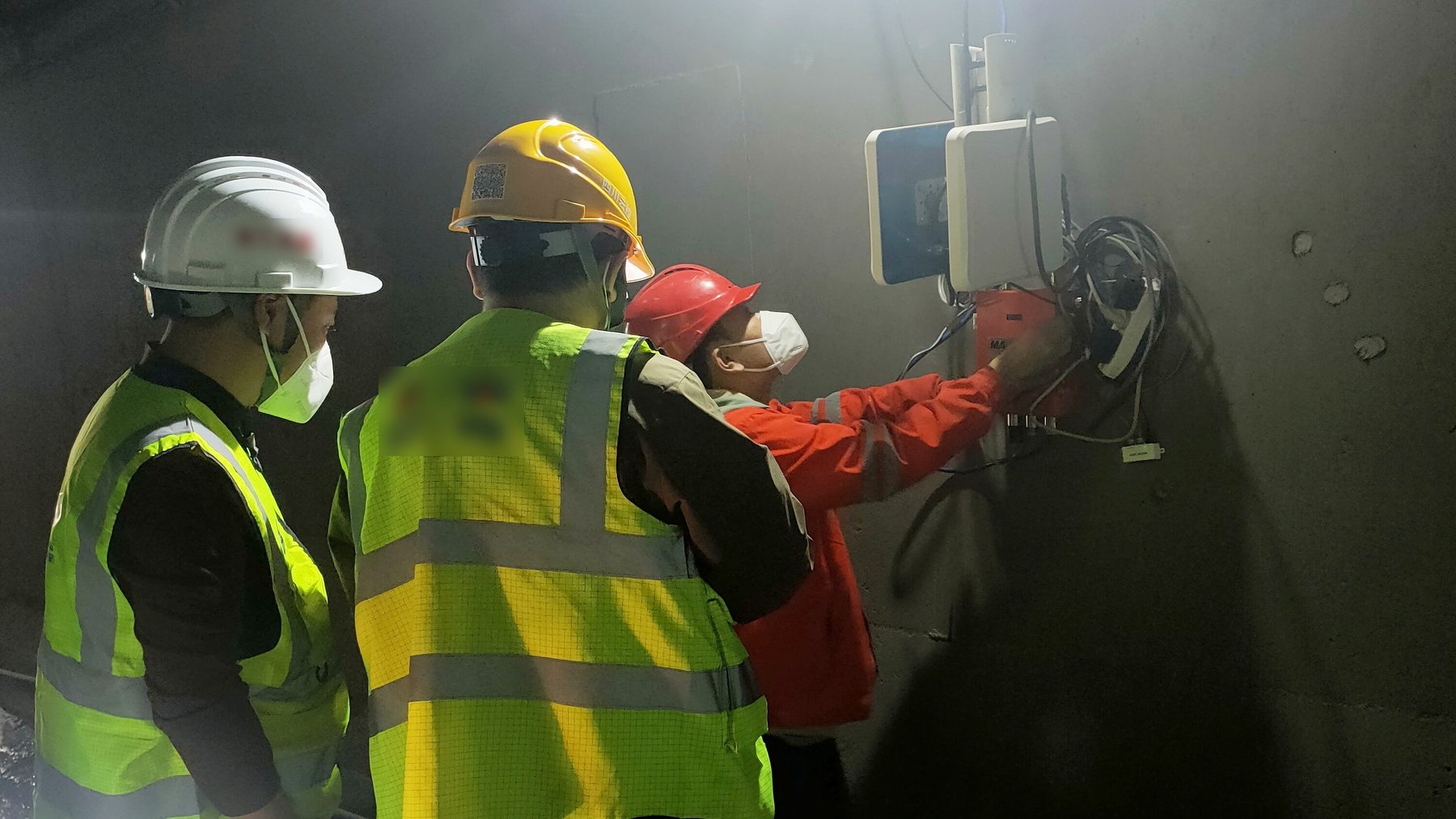 Three workers in hard hats and high-visibility vests installing a UWB anchor on the wall of a highway tunnel under construction